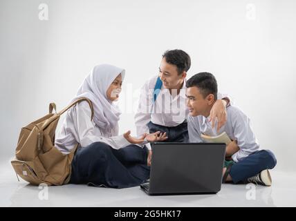 tre adolescenti in divise junior della scuola superiore che chiacchierano quando si siedono sul pavimento mentre si usa un computer portatile insieme Foto Stock