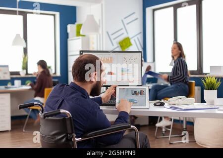 Lavoratore non valido minorato seduto immobilizzato in sedia a rotelle lavorando al blocco note e computer contemporaneamente analizzando grafici finanziari, pc che mostra la prosessione di dati per il progetto riguardante l'economia. Foto Stock