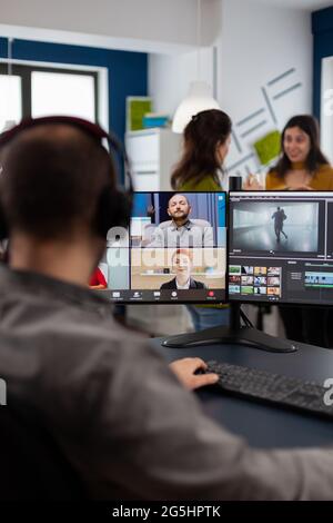 Dipendente videografo che ha una videoconferenza indossando cuffie per l'editing di filmati client, ottenendo feedback sul progetto commerciale. Freelance che utilizza software di post-produzione su pc in ufficio creativo. Foto Stock