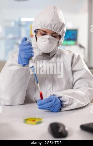 Scienziato vestito con tuta ppe che preleva il campione di sangue utilizzando una micropipetta. Scienziato vestito in tuta ppe prelievo campione di sangue chimico in tuta lavorando con vari batteri, campioni di sangue di tessuto per la ricerca di antibiotici. Foto Stock