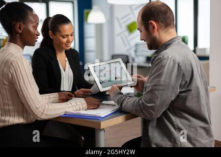 Entusiasta uomini d'affari diversi leggere il rapporto finanziario annuale seduta al tavolo in un moderno ufficio aziendale startup tenendo tablet e sorridendo. Team di imprenditori multietnici che lavorano in azienda Foto Stock