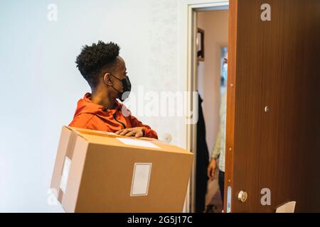 Giovane addetto all'assistenza essenziale che consegna il pacchetto a un cliente maschile a casa durante il COVID-19 Foto Stock