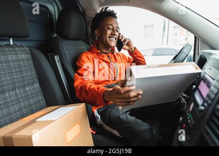 Sorridente addetto all'assistenza essenziale che guarda il pacchetto mentre parla con lo smartphone in un pulmino di consegna Foto Stock