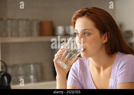 Donna seria che beve acqua dal vetro in cucina a casa Foto Stock