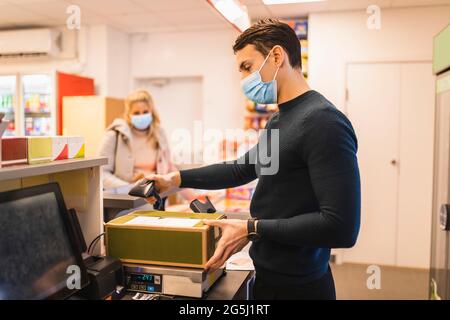 Imprenditore maschile che utilizza il lettore di codici a barre sul pacchetto mentre la cliente femminile si trova al momento del pagamento durante la pandemia Foto Stock
