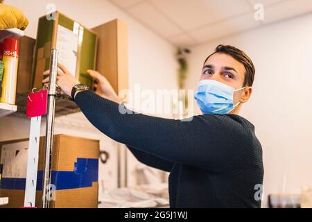 Il proprietario del negozio maschile indossa una maschera protettiva mentre rimuove la scatola dallo scaffale Foto Stock