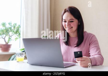 Una giovane donna tiene una carta di credito in mano e fa un pagamento online su Internet - UNA ragazza utilizza un computer portatile e una carta per lo shopping online - il Foto Stock