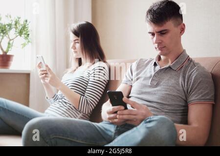 I giovani offesi sono seduti a casa sul divano e guardando il telefono - UNA coppia litigata e seduta separatamente - UN rapporto rovinato tra Foto Stock