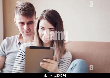 Il giovane abbraccio dolcemente la sua ragazza e sua moglie e guarda il contenuto sul tablet - giovane coppia si rilassa sul divano e fa acquisti in linea Foto Stock