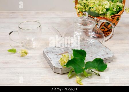 Teiera vuota in vetro e due tazze per il tè su un tavolo di legno sullo sfondo di foglie di lime e fiori. Tè healing. Foto Stock