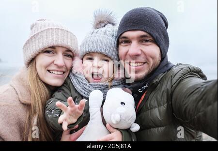 La famiglia felice prende un selfie - i genitori ed il bambino scattano le foto di se stessi sul telefono - papà, mamma e figlia hanno divertimento davanti alla macchina fotografica del telefono Foto Stock