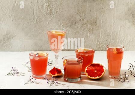 Un mazzo di bicchieri, bicchieri con diversi cocktail, bevande e fiori su uno sfondo di cemento con ombre dure Foto Stock