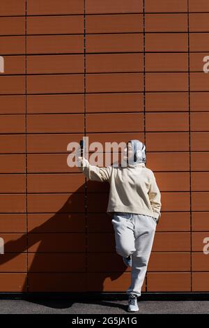 Ritratto grafico a lunghezza intera di sicura donna mediorientale che prende selfie in città strada mentre si sta in piedi da muro di mattoni, copia spazio Foto Stock