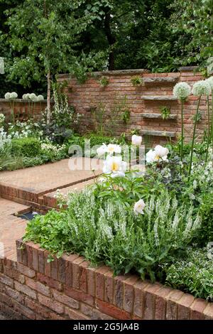 Patio in mattoni a spina di pesce, pareti in mattoni rigenerati, acqua rossiccia, cubetti e piantine bianche, aquilegia, allidi, gerani e rose Foto Stock