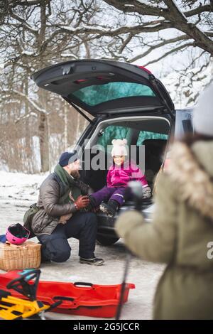 Tutta la lunghezza del padre che aiuta la figlia che indossa la scarpa seduta nel bagagliaio dell'automobile durante l'inverno Foto Stock