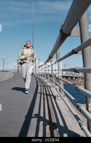 Ritratto verticale a lunghezza intera della moderna donna mediorientale che corre verso la fotocamera sul lungofiume illuminato dalla luce del sole Foto Stock