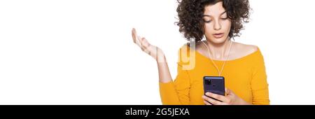Scioccata giovane ragazza arrabbiata che guarda il suo cellulare in incredulità. Adolescente fissando un messaggio di testo scioccante sul suo telefono, isolato su banner bianco. Foto Stock