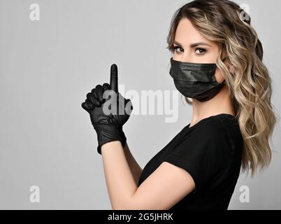 Ritratto di donna riccia in t-shirt nera, maschera e guanti in lattice in piedi lateralmente tenendo le mani insieme, pistola dito Foto Stock