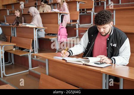 Uno degli studenti che utilizza lo smartphone mentre prende appunti durante la lezione Foto Stock