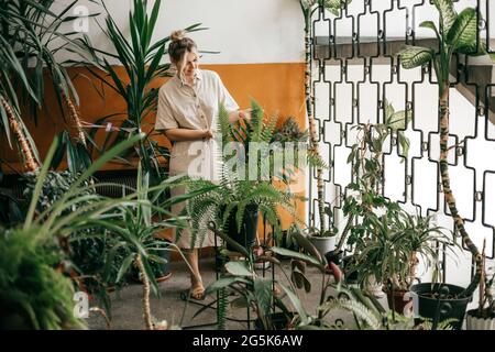 Foto di una giovane donna a tutta lunghezza in abiti leggeri fatti di tessuto naturale in una piccola sala formale di un bel palazzo d'ufficio, piantagione di piante Foto Stock