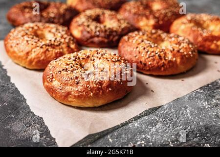 Primo piano appetitosi e croccanti bagel con semi di sesamo e lino. Cotto caldo fatto in casa. Bagel su carta da forno su fondo grigio di cemento. Cuocere alle ore Foto Stock