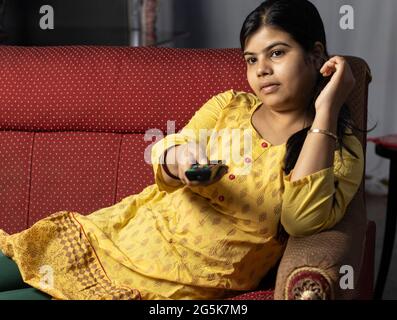 Una casalinga indiana che guarda la televisione tenendo il telecomando in mano, seduto sul divano Foto Stock