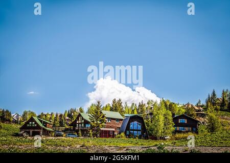 Gruppo di capanne di tronchi e case in collina con cime innevate in lontananza e pini e aspen Foto Stock