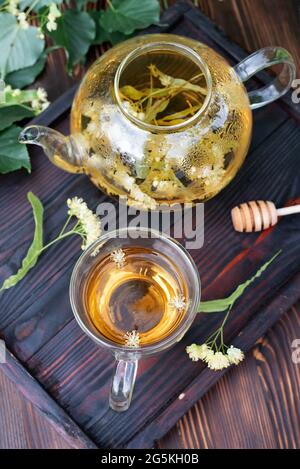 Teiera di vetro e una tazza di tè tiglio su un vassoio di legno scuro sul tavolo, sano tè alle erbe, primo piano. Foto Stock