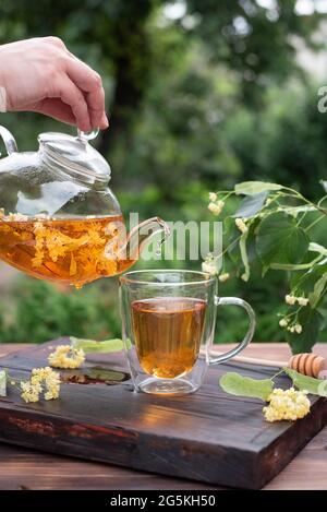 Tè sano alle erbe, mani femminili che versano il tè tiglio da una teiera di vetro in una tazza nel giardino, primo piano. Foto Stock