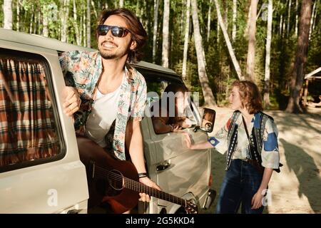 Giovane uomo in occhiali da sole con chitarra in viaggio con i suoi amici nella foresta Foto Stock