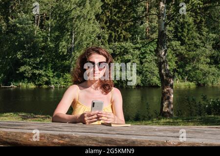 Una ragazza con gli occhiali tiene uno smartphone in mano, una ragazza si siede a un tavolo di legno. Attività ricreative e di intrattenimento fuori città. Foto Stock