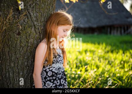 Ragazza giovane con capelli rossi in piedi da albero, fienile in background. Foto Stock