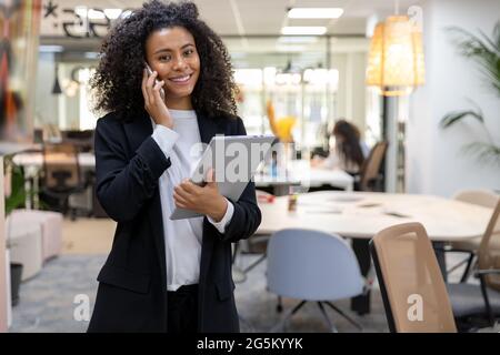 Felice donna d'affari africana che parla al telefono sorridendo e guardando la macchina fotografica. Foto Stock