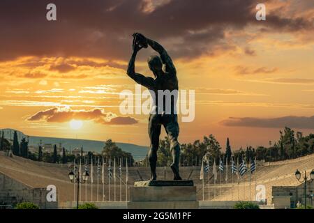 La statua in bronzo del discobolo, l'antico stadio Panathinaiko sullo sfondo del tramonto, scultura in metallo diskovolos vicino a Kallimarmaro Atene. Grecia. Foto Stock