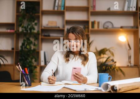 Sorridente giovane imprenditore attraente che usa il telefono cellulare mentre lavora con documenti alla scrivania dell'ufficio Foto Stock