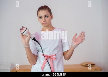 Attraente giovane medico possiede un apparecchio di correzione della figura. Terapista massaggiante femminile che fa la procedura di cavitazione. Cosmetologia a radiofrequenza Foto Stock