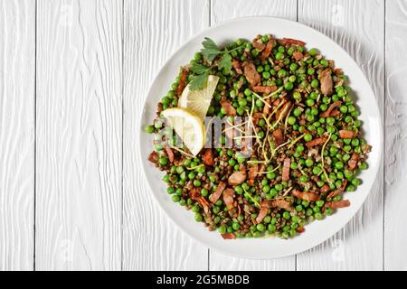 Insalata libanese di lenticchie calde e piselli verdi con pancetta, scorza di limone e condimento di limone e olio d'oliva servito su un piatto bianco su uno sfondo di legno bianco Foto Stock