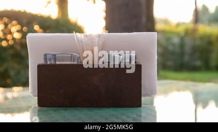 Un porta tovaglioli di legno marrone e stuzzicadenti siedono sul tavolo di vetro del caffè, illuminato dai raggi del sole. Tavolo nel ristorante. Vetro Foto Stock