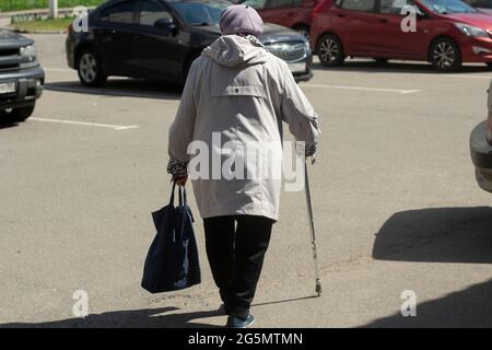 Un pensionato con un bastone sta camminando lungo la strada. Una donna anziana con una gamba irritata si appoggia su una canna. Nonna va al negozio. Foto Stock