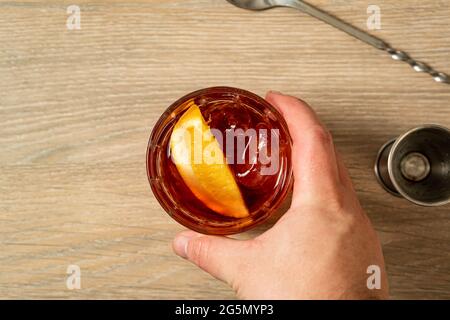 Vista dall'alto del cocktail negroni che viene afferrato con una mano con cucchiaio e jigger sulla superficie di legno. Foto Stock