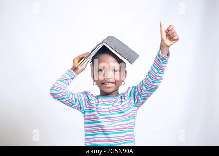 Carino bambino africano ragazza con stile dei capelli intrecciati mette felicemente un libro sulla sua testa, mentre studiando per mostrare come interessante educazione è diventata Foto Stock