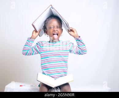 Carino bambino africano ragazza con stile dei capelli intrecciati mette felicemente un libro sulla sua testa, mentre studiando per mostrare come interessante educazione è diventata Foto Stock