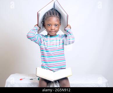 Carino bambino africano ragazza con stile dei capelli intrecciati mette felicemente un libro sulla sua testa, mentre studiando per mostrare come interessante educazione è diventata Foto Stock