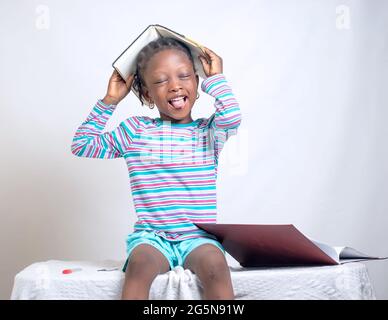 Carino bambino africano ragazza con stile dei capelli intrecciati mette felicemente un libro sulla sua testa, mentre studiando per mostrare come interessante educazione è diventata Foto Stock