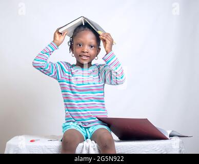 Carino bambino africano ragazza con stile dei capelli intrecciati mette felicemente un libro sulla sua testa, mentre studiando per mostrare come interessante educazione è diventata Foto Stock