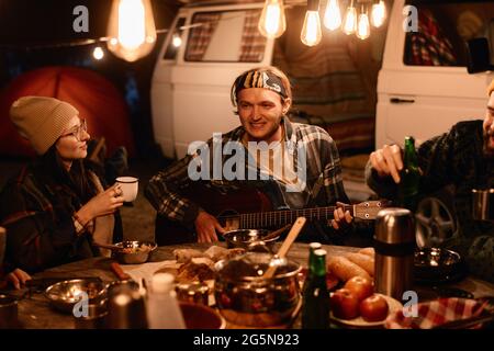 Giovane che suona la chitarra seduto al tavolo con i suoi amici e cena la sera in un picnic Foto Stock