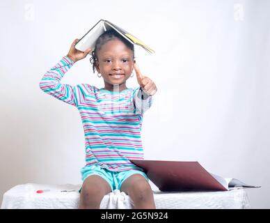 Carino bambino africano ragazza con stile dei capelli intrecciati mette felicemente un libro sulla sua testa, mentre studiando per mostrare come interessante educazione è diventata Foto Stock