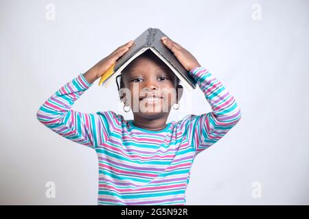 Carino bambino africano ragazza con stile dei capelli intrecciati mette felicemente un libro sulla sua testa, mentre studiando per mostrare come interessante educazione è diventata Foto Stock