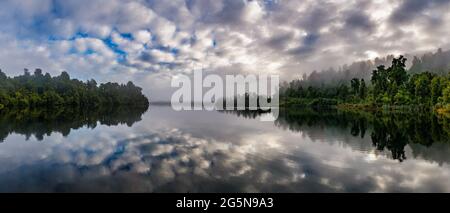 Riflessi nuvolosi su una mattina misteriosa di un cespuglio e foresta fiancheggiata Lago Mapourika nella Costa Ovest della Nuova Zelanda Foto Stock