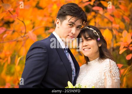 Sposa biraciale e sposo insieme all'aperto dopo il matrimonio autunnale con brillanti foglie arancioni colorate sullo sfondo Foto Stock